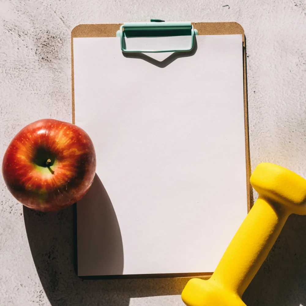 Zdjęcie ukazuje betonową podłogę, a na niej notes z zieloną klamerką, jabłko oraz żółty mały ciężarek. Całość oświetla słońce, a przedmioty rzucają cień.