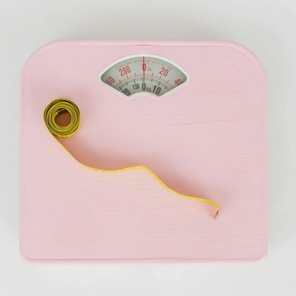 Różowa waga łazienkowa z chropowatą powierzchnią. Na niej leży żółta metrówka krawiecka.