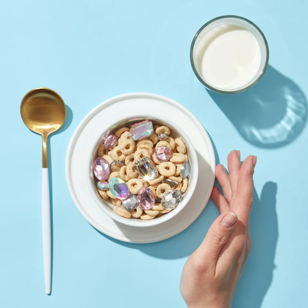 Miska z płatkami śniadaniowymi i brylancikami, obok złota łyżka i szklanka z mlekiem. Wszystkie przedmioty stoją na niebieskim tle.